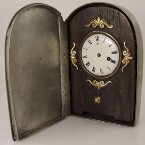 May include: An antique clock with a dark wooden face and a metal casing. The clock face is white with black Roman numerals and gold-colored decorative accents. The metal casing has a hinged door that is open.