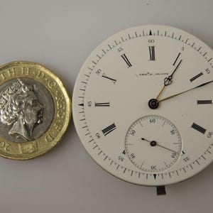 May include: A white watch face with black Roman numerals and hands, alongside a &pound;1 coin. The watch face has a smaller seconds dial and the text "CHAS. E. JAESCH" printed on it. The coin is dated 2018.