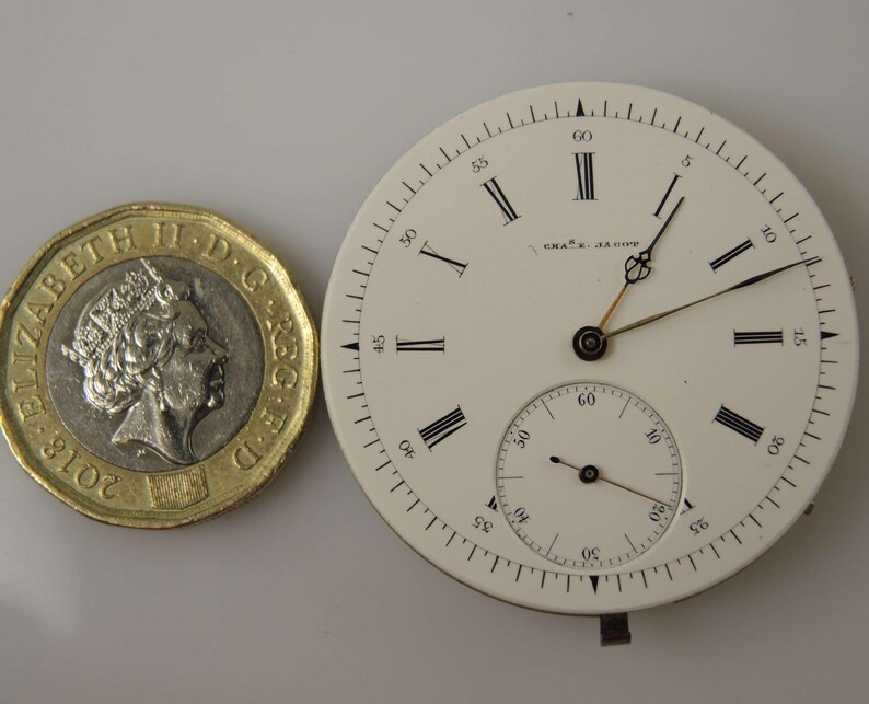 May include: A white watch face with black Roman numerals and hands, alongside a &pound;1 coin. The watch face has a smaller seconds dial and the text "CHAS. E. JAESCH" printed on it. The coin is dated 2018.
