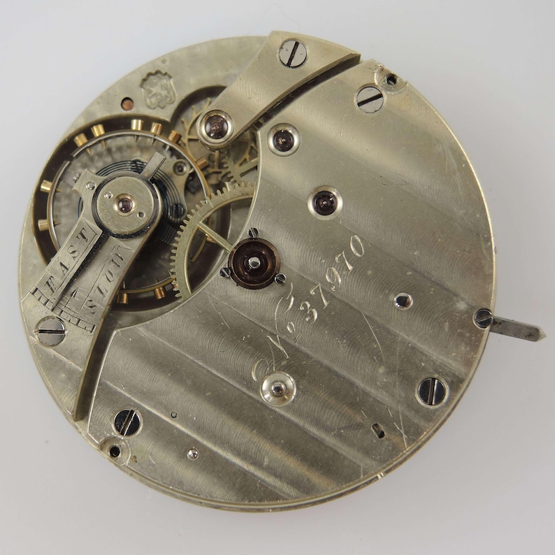 May include: Close-up of a vintage watch movement. The silver-toned mechanism features intricate gears, screws, and a balance wheel. The dial reads "FAST" and "SLOW". The serial number "No. 37970" is engraved on the surface. A small lever extends from the side.