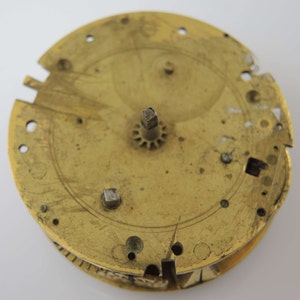 May include: A brass clock face with a central gear and several holes around the edge. The face is worn and has a patina.
