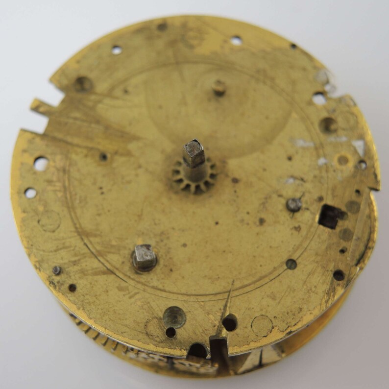 May include: A brass clock face with a central gear and several holes around the edge. The face is worn and has a patina.