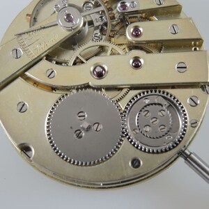 Puede incluir: Primer plano de un movimiento de reloj vintage con engranajes y tornillos intrincados. El movimiento es de color dorado y tiene un engranaje plateado en el centro. El movimiento del reloj se est&aacute; trabajando con un peque&ntilde;o destornillador.