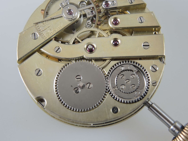 Puede incluir: Primer plano de un movimiento de reloj vintage con engranajes y tornillos intrincados. El movimiento es de color dorado y tiene un engranaje plateado en el centro. El movimiento del reloj se est&aacute; trabajando con un peque&ntilde;o destornillador.