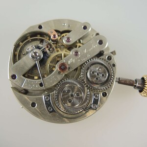Op de afbeelding: Een zilveren uurwerk met ingewikkelde tandwielen en een opwindslinger. Het uurwerk is gesigneerd met "Alfred  Michel & Cie.  La Chaux-de-Fonds."