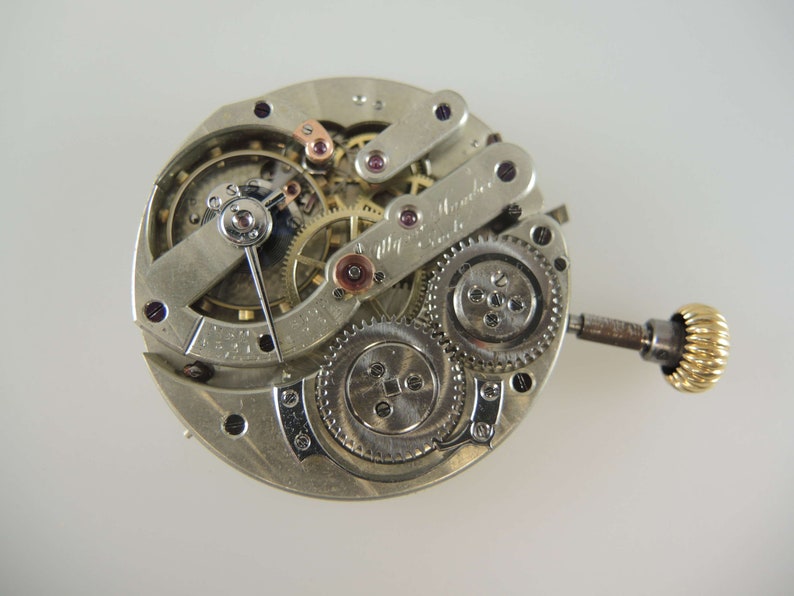 Op de afbeelding: Een zilveren uurwerk met ingewikkelde tandwielen en een opwindslinger. Het uurwerk is gesigneerd met "Alfred  Michel & Cie.  La Chaux-de-Fonds."