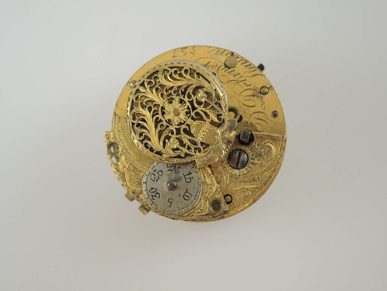 K&ouml;nnte beinhalten: Ein goldfarbenes Taschenuhrwerk mit aufwendigen floralen Gravuren. Das Zifferblatt der Uhr ist mit r&ouml;mischen Ziffern und einem kleinen, kreisf&ouml;rmigen Zifferblatt mit den Zahlen 5, 10, 15, 20, 25, 30 sichtbar.