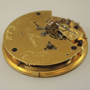 May include: A gold pocket watch with the inscription "James Liverpool" on the back. The watch is open, revealing the intricate inner workings of the movement.