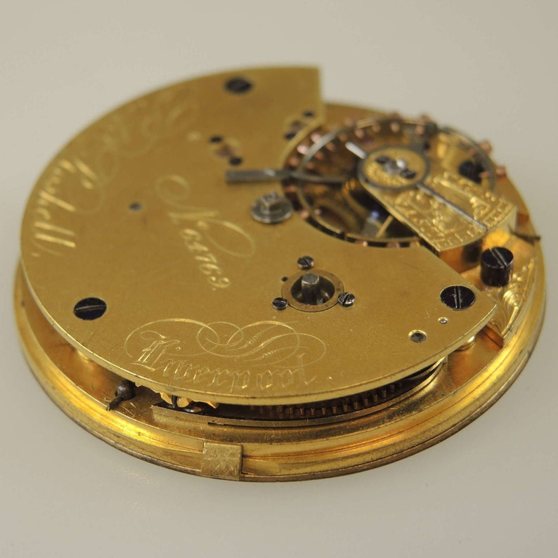 May include: A gold pocket watch with the inscription "James Liverpool" on the back. The watch is open, revealing the intricate inner workings of the movement.