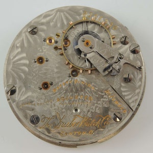 May include: Close-up of the back of a silver pocket watch with a gold-colored inscription that reads "The Huber Watch Co. Canton, O." and "Safety Pinion Adjusted For Five Positions."