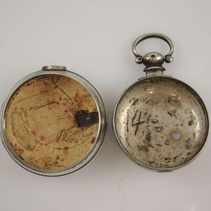 Könnte beinhalten: Das Innere einer silbernen Taschenuhr mit einem Scharnierdeckel. Der Deckel ist geöffnet und zeigt ein Papieretikett mit Schrift. Das Zifferblatt der Uhr ist geschlossen und trägt die Nummer "14".