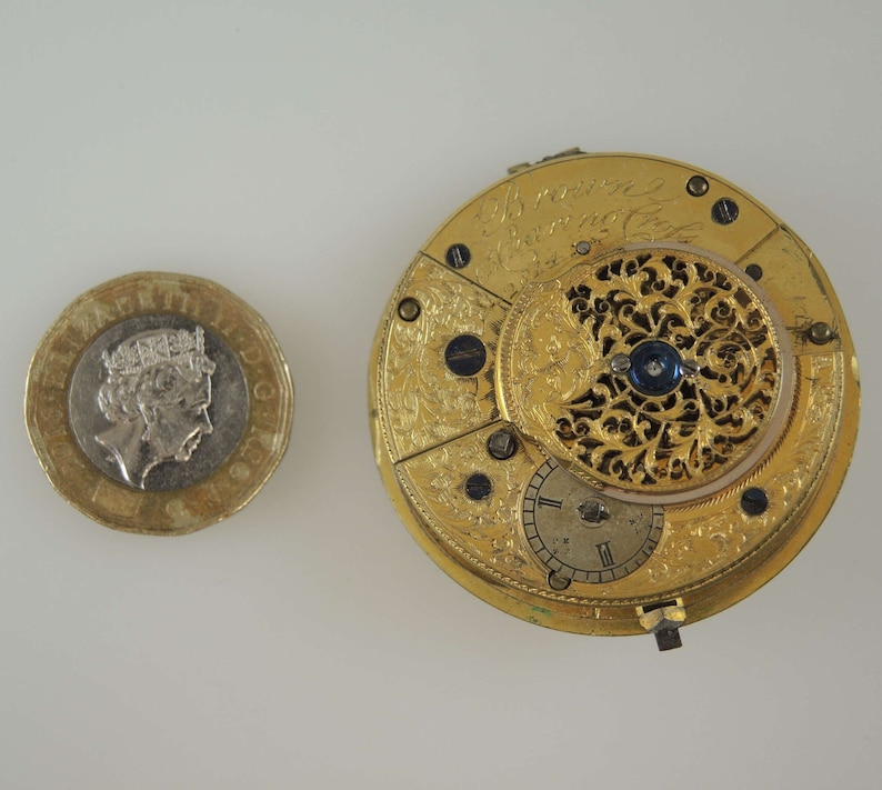 Puede incluir: Un reloj de bolsillo de oro con grabados intrincados y un centro azul. La esfera del reloj es visible y tiene marcas de n&uacute;meros romanos.