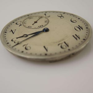 K&ouml;nnte beinhalten: Eine silberne Taschenuhr mit schwarzen r&ouml;mischen Ziffern f&uuml;r die Stunden und schwarzen Zeigern. Das Zifferblatt der Uhr hat ein kleines Hilfszifferblatt mit schwarzen arabischen Ziffern f&uuml;r die Minuten.