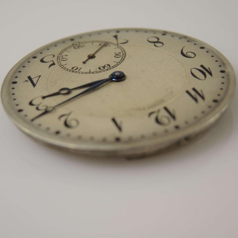 K&ouml;nnte beinhalten: Eine silberne Taschenuhr mit schwarzen r&ouml;mischen Ziffern f&uuml;r die Stunden und schwarzen Zeigern. Das Zifferblatt der Uhr hat ein kleines Hilfszifferblatt mit schwarzen arabischen Ziffern f&uuml;r die Minuten.