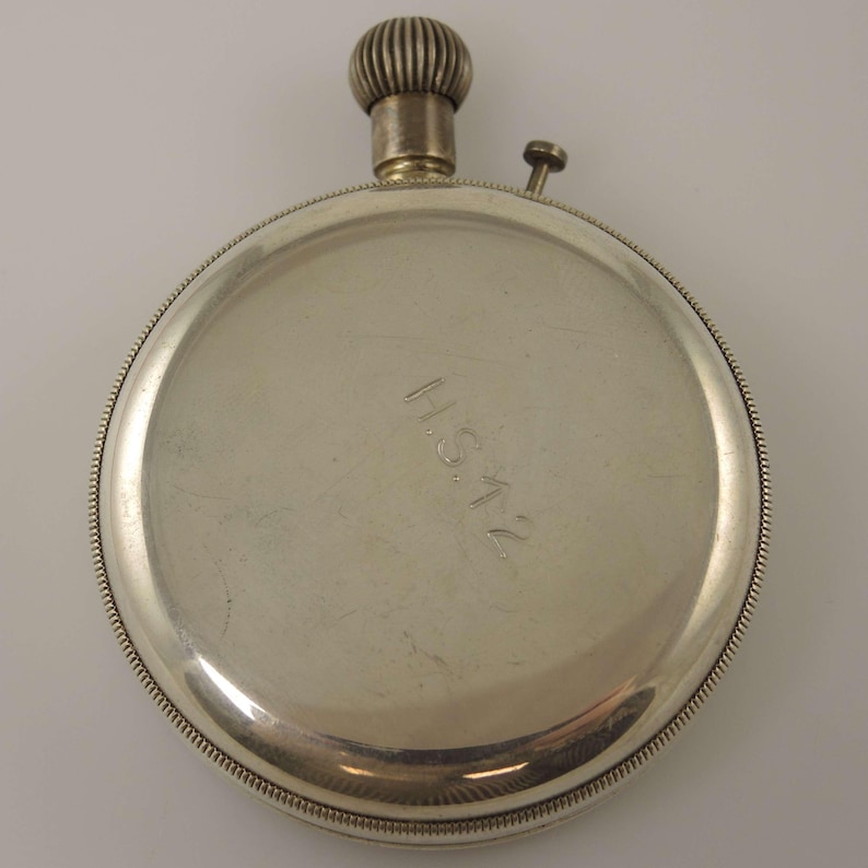 K&ouml;nnte beinhalten: Eine silberfarbene Taschenuhr mit einer strukturierten Aufzugskrone und einem kleinen Knopf. Die Uhr hat ein kreisf&ouml;rmiges Zifferblatt mit einem Perlenrand und den eingravierten Buchstaben "H.S.12".