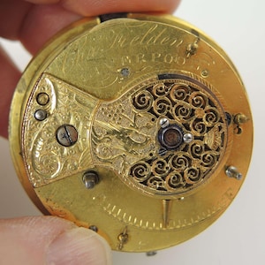 May include: Close-up of a vintage gold pocket watch movement. The watch mechanism features intricate engraved details, including floral patterns and the name "Fielden" above "Liverpool". The watch has a circular design with various screws and gears.
