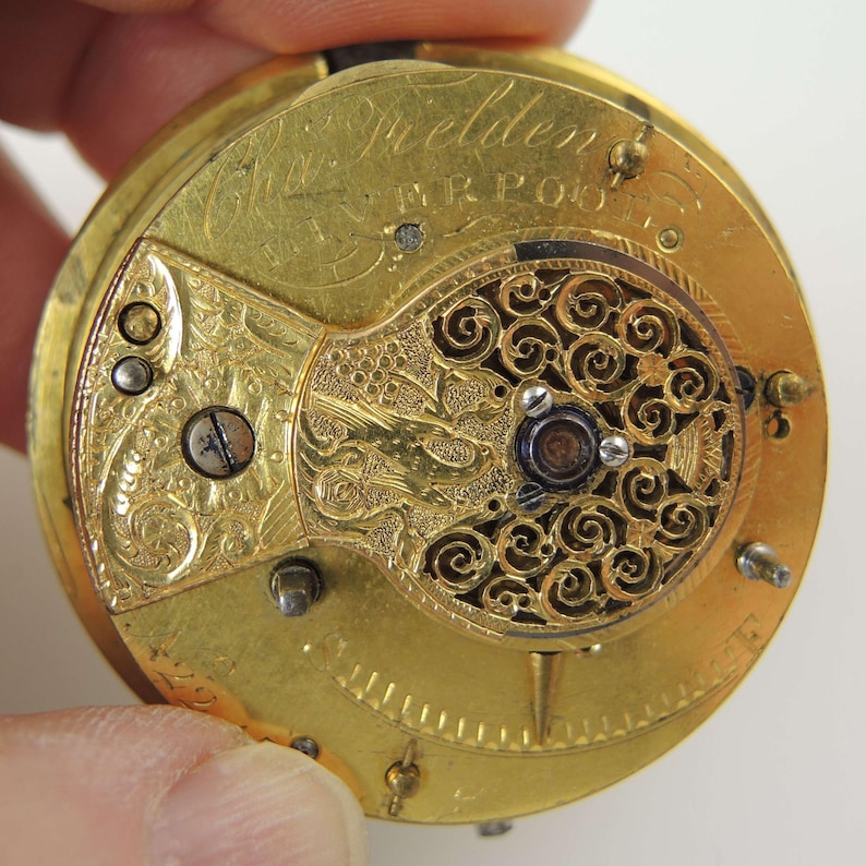 May include: Close-up of a vintage gold pocket watch movement. The watch mechanism features intricate engraved details, including floral patterns and the name "Fielden" above "Liverpool". The watch has a circular design with various screws and gears.