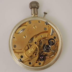 K&ouml;nnte beinhalten: Nahaufnahme des Uhrwerks einer antiken Taschenuhr. Die Uhr hat ein silberfarbenes Geh&auml;use und ein goldfarbenes Innenwerk mit komplizierten Zahnr&auml;dern, Schrauben und Juwelen. Oben sind eine silberne Krone und ein kleiner Knopf zu sehen. Wei&szlig;er Hintergrund.