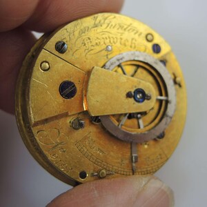 May include: Close-up of a vintage watch movement with gold-colored gears and components. The watch mechanism has engraved text, including the name "Wm Gurton Norwich" and the number "9141". The intricate design features small screws and a balance wheel.