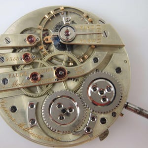 Puede incluir: Primer plano de un movimiento de reloj de bolsillo plateado con la inscripci&oacute;n "Baume & Mercier" y "Trist Meubles" en la esfera. El reloj tiene un acabado plateado y engranajes intrincados.