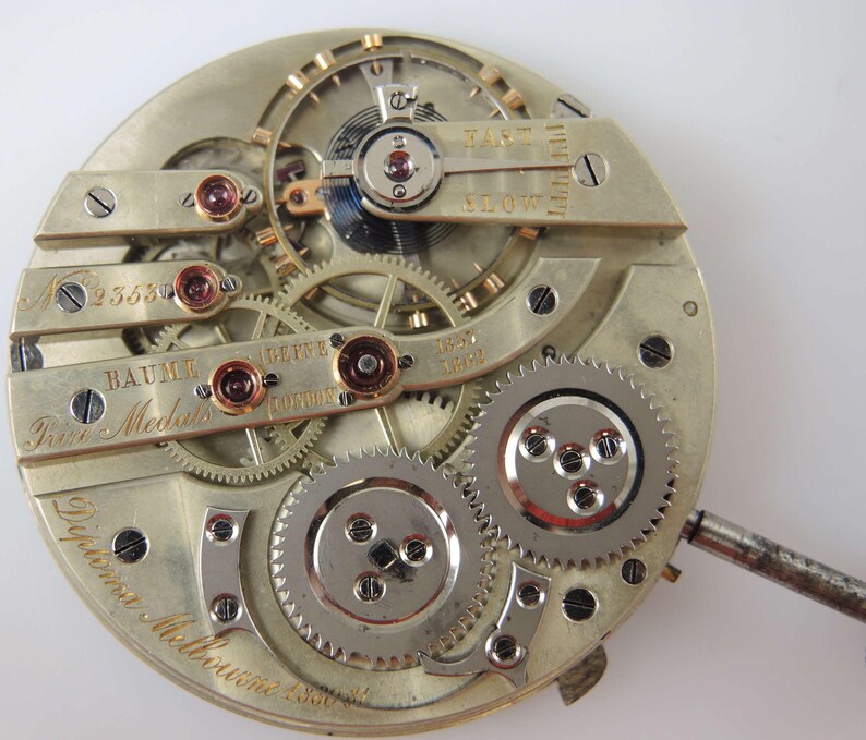 Puede incluir: Primer plano de un movimiento de reloj de bolsillo plateado con la inscripci&oacute;n "Baume & Mercier" y "Trist Meubles" en la esfera. El reloj tiene un acabado plateado y engranajes intrincados.