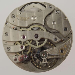 May include: Close-up of a vintage watch movement with intricate gears, screws, and ruby jewels. The silver-toned mechanism features the text "21 Jewels" and "Swiss Made". A detailed view of the inner workings of a mechanical timepiece.