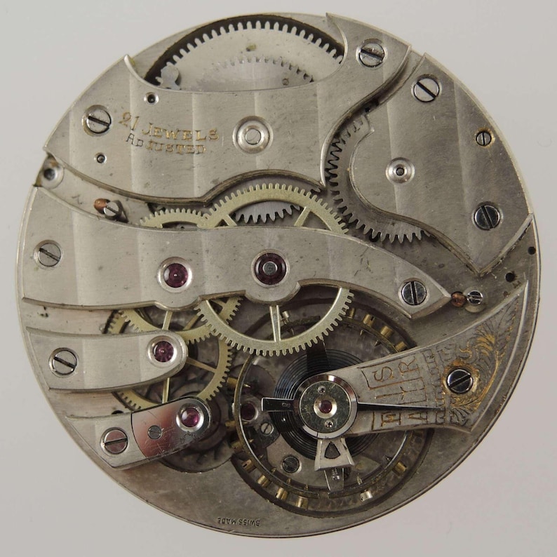 May include: Close-up of a vintage watch movement with intricate gears, screws, and ruby jewels. The silver-toned mechanism features the text "21 Jewels" and "Swiss Made". A detailed view of the inner workings of a mechanical timepiece.