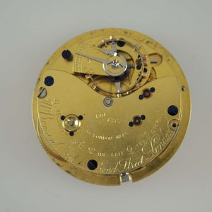 Op de afbeelding: Een close-up van een goudkleurig uurwerk met ingewikkelde tandwielen, schroeven en een balanswiel. Het uurwerk heeft tekst die luidt "178 Bond Street London". Dit is een vintage horloge.