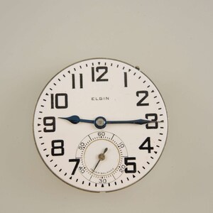 Op de afbeelding: Een wit horloge-wijzerplaat met zwarte cijfers en wijzers. Op de wijzerplaat staat bovenaan in het zwart "ELGIN" gedrukt. De wijzerplaat is omgeven door een zilverkleurige rand.
