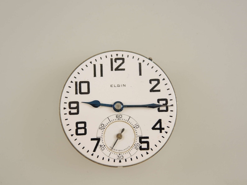 Op de afbeelding: Een wit horloge-wijzerplaat met zwarte cijfers en wijzers. Op de wijzerplaat staat bovenaan in het zwart "ELGIN" gedrukt. De wijzerplaat is omgeven door een zilverkleurige rand.