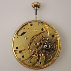 May include: A gold-colored pocket watch movement with intricate gears and a winding stem. The movement is labeled "Moebius Werke" and has the serial number "6D0648".