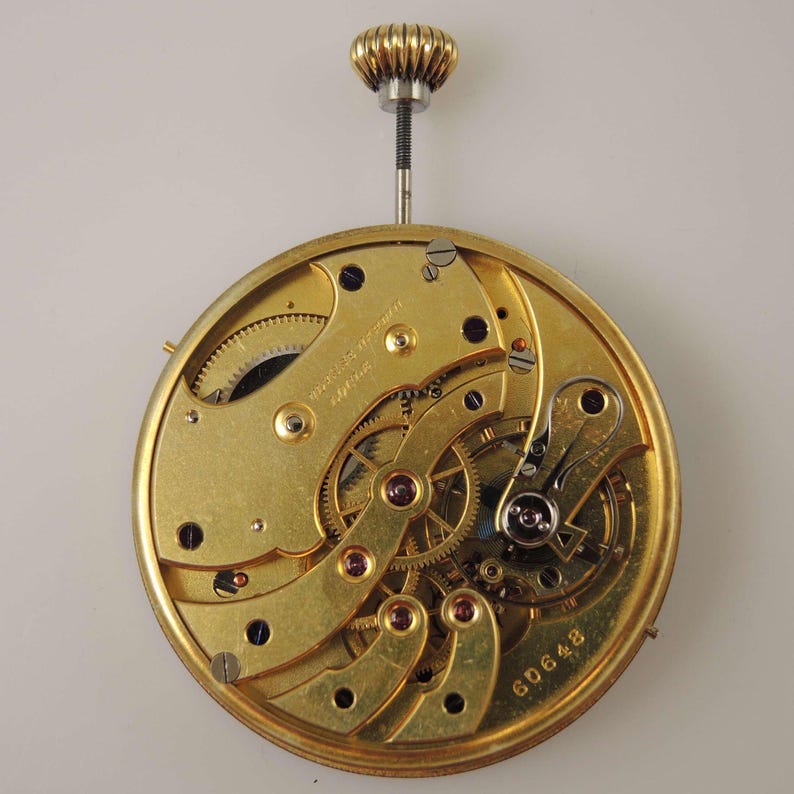 May include: A gold-colored pocket watch movement with intricate gears and a winding stem. The movement is labeled "Moebius Werke" and has the serial number "6D0648".