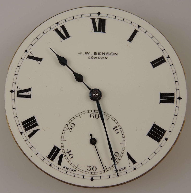 Puede incluir: Una esfera de reloj de bolsillo blanca con marcadores de hora en n&uacute;meros romanos negros y agujas negras. La esfera del reloj tambi&eacute;n tiene un c&iacute;rculo conc&eacute;ntrico m&aacute;s peque&ntilde;o con marcas negras para los segundos. La esfera del reloj est&aacute; etiquetada como "J.W. Benson London" y "Swiss Made".