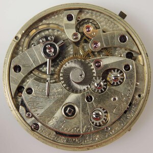 May include: Detailed view of a vintage pocket watch mechanism. The silver-coloured movement displays intricate gears, screws, and a balance wheel. The words "Fast Slow" are visible, along with the name "The E. Jacobs" and patent details.