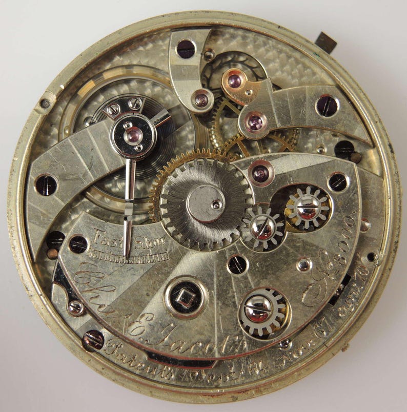 May include: Detailed view of a vintage pocket watch mechanism. The silver-coloured movement displays intricate gears, screws, and a balance wheel. The words "Fast Slow" are visible, along with the name "The E. Jacobs" and patent details.