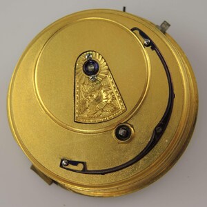 Pode incluir: Close-up do mecanismo de um rel&oacute;gio de bolso dourado. O rel&oacute;gio apresenta detalhes intrincados, incluindo um escudo decorativo com padr&otilde;es florais e de folhas, e um componente curvo azul escuro. Uma pequena gema azul escura est&aacute; definida no topo do escudo.