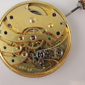 May include: Close-up of a gold-toned watch movement with intricate gears and a winding stem. The movement is labeled "W.B. Watch Co. London" and has the number "6D648" inscribed on the bottom.