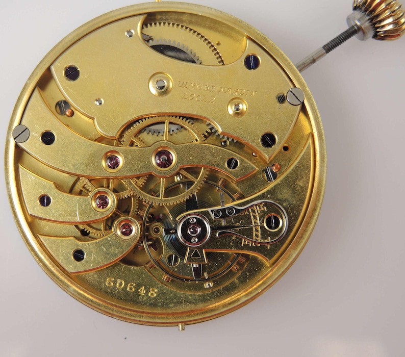 May include: Close-up of a gold-toned watch movement with intricate gears and a winding stem. The movement is labeled "W.B. Watch Co. London" and has the number "6D648" inscribed on the bottom.