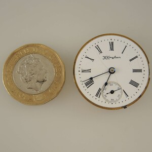 Puede incluir: Una esfera de reloj de bolsillo blanca con marcadores de hora en n&uacute;meros romanos negros y agujas negras. La esfera del reloj tiene el texto "J. Hampden" impreso en ella. La esfera del reloj est&aacute; rodeada por una carcasa de metal de color dorado.