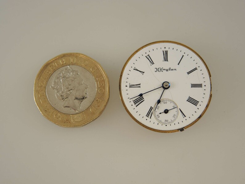 Puede incluir: Una esfera de reloj de bolsillo blanca con marcadores de hora en n&uacute;meros romanos negros y agujas negras. La esfera del reloj tiene el texto "J. Hampden" impreso en ella. La esfera del reloj est&aacute; rodeada por una carcasa de metal de color dorado.