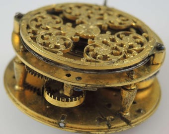 May include: A gold-toned pocket watch with intricate openwork design on the back. The watch face is not visible. The watch is disassembled, showing the inner workings of the mechanism.