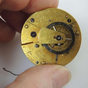 May include: Close-up of a vintage watch movement with a gold-colored finish. The mechanism features intricate gears, screws, and a balance wheel. The text "Slow" and "Fast" are visible, along with the number "19191".