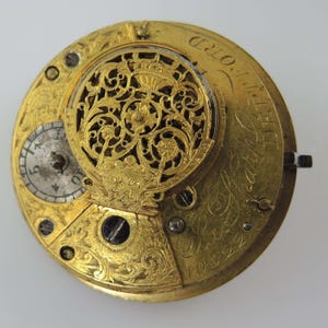Puede incluir: Primer plano de un movimiento de reloj dorado antiguo. El intrincado mecanismo presenta filigranas ornamentadas, una pequeña esfera y texto grabado. Los componentes del reloj están asegurados con pequeños tornillos, mostrando la artesanía del reloj.