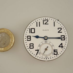 Op de afbeelding: Een wit zakhorloge met zwarte cijfers en wijzers. Het horloge heeft de tekst "ELGIN" op de wijzerplaat en een klein sub-wijzerplaat met zwarte markeringen.