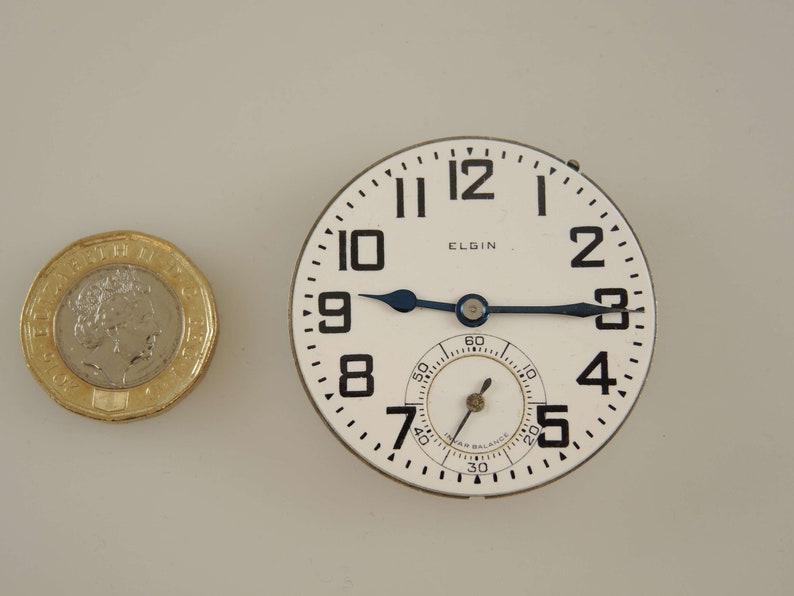 Op de afbeelding: Een wit zakhorloge met zwarte cijfers en wijzers. Het horloge heeft de tekst "ELGIN" op de wijzerplaat en een klein sub-wijzerplaat met zwarte markeringen.