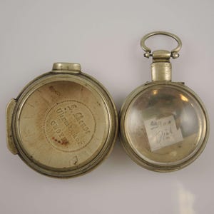 Könnte beinhalten: Zwei antike silberne Taschenuhren, eine mit einem klaren Glaszifferblatt und einer handschriftlichen Notiz im Inneren, die andere mit einem silbernen Zifferblatt und der Inschrift "J. Allen, Watchmaker, Chester" auf der Rückseite.