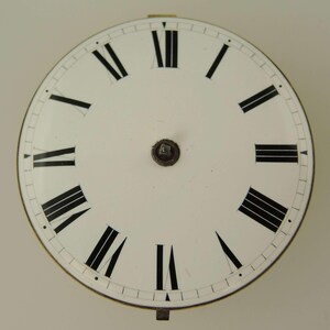 May include: A white clock face with black Roman numerals marking the hours. The face has a metal rim and a small central mechanism. The hands are missing. The clock face is a vintage or antique item.