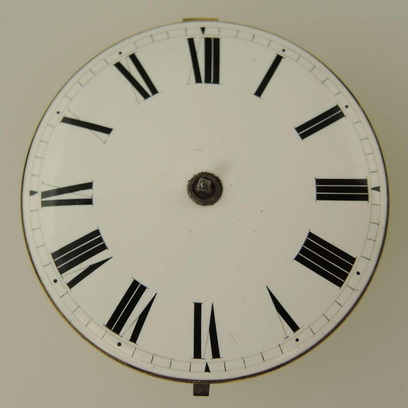 May include: A white clock face with black Roman numerals marking the hours. The face has a metal rim and a small central mechanism. The hands are missing. The clock face is a vintage or antique item.