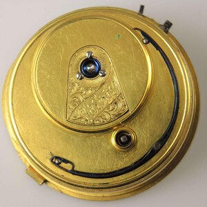 May include: Close-up of a gold-colored pocket watch mechanism. The watch features intricate engraved details and a blue jewel at the center. A black curved line and small screws are also visible.
