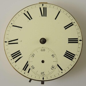 May include: Antique watch face with a cream-colored background and black Roman numerals. A smaller dial for seconds is at the bottom. The watch face has a gold-colored metal rim and a small, decorative knob at the center.